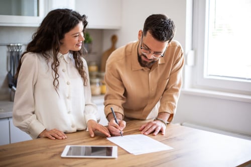 Deux personnes discutent et rédigent un bail de location dans une cuisine moderne et lumineuse. Un homme portant des lunettes et une chemise beige remplit attentivement un document papier avec un stylo, tandis qu’une femme en chemise blanche l’observe avec intérêt, assise à ses côtés devant une table en bois. Un ordinateur portable ouvert est posé sur la table, suggérant une vérification ou une comparaison des clauses du contrat. Cette scène illustre les précautions à prendre lors de la rédaction d’un bail pour éviter les erreurs courantes et sécuriser la relation entre propriétaire et locataire. Idéal pour accompagner un article sur les bonnes pratiques en matière de rédaction de bail locatif.