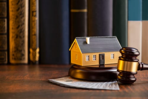 Petit modèle de maison jaune posé sur un bureau en bois devant un marteau de juge et des billets de banque symbolisant un litige immobilier. Cette image illustre les risques de surestimation immobilière pouvant mener à des poursuites judiciaires pour un agent immobilier. Contexte juridique et financier lié aux conflits entre vendeurs, acheteurs et professionnels de l'immobilier lors d'une évaluation erronée de la valeur d'un bien. Idéale pour un article sur les conséquences légales et les recours en cas de surévaluation immobilière.