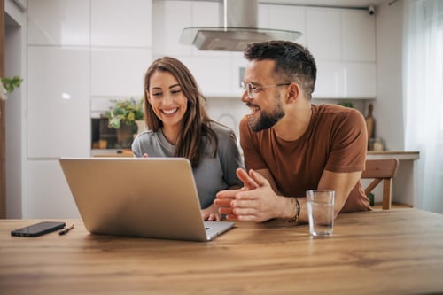 Jeune couple souriant consultant des informations sur un ordinateur portable dans une cuisine moderne et lumineuse. Les deux personnes semblent discuter avec enthousiasme des démarches administratives liées aux aides au logement. L’homme et la femme, assis à une table en bois, examinent ensemble des données en ligne, probablement pour vérifier leur éligibilité aux aides financières comme les APL ou autres dispositifs d’aide au logement en 2026. Ambiance conviviale et studieuse reflétant la recherche d’informations pratiques pour les locataires. Contexte adapté à un article sur les critères d’éligibilité aux aides au logement.