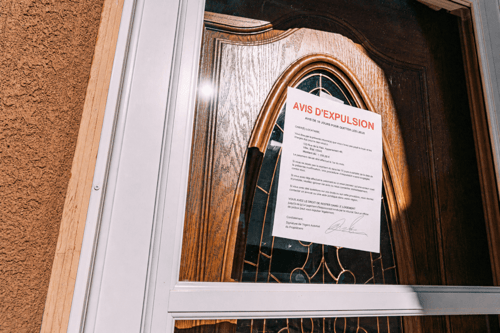 Avis d’expulsion locative affiché sur une porte en bois ancienne d’un logement en France. Ce document officiel, intitulé AVIS D’EXPULSION, indique un délai de quelques jours pour quitter les lieux sous peine de recours juridique. Il est apposé sur une porte intérieure à panneaux moulurés avec une vitre décorative et encadrement en bois sculpté, reflétant un habitat potentiellement ancien ou de caractère. Le texte précise les conséquences légales en cas de non-respect du délai et mentionne l’intervention possible d’un huissier. Cette image illustre concrètement les procédures d’expulsion locative en débat dans l’article sur l’inapplicabilité des arrêtés municipaux anti-expulsions, mettant en lumière les tensions entre régulations locales et cadre juridique national en matière de logement.