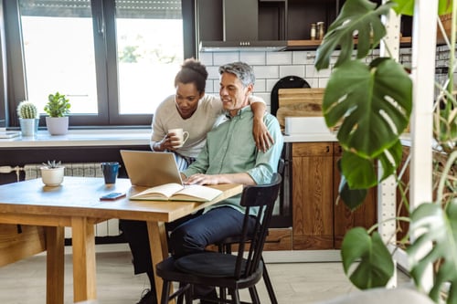 Deux personnes discutent des options d’assurance habitation pour locataires dans une cuisine moderne et lumineuse. L’homme, assis à une table en bois avec un ordinateur portable ouvert devant lui, consulte des documents tout en souriant. La femme, debout à ses côtés, pointe un détail sur l’écran tout en tenant une tasse de café. La pièce est décorée avec des plantes vertes, des pots sur le rebord de la fenêtre et une étagère en bois, créant une ambiance chaleureuse et organisée. Cette scène illustre la recherche d’une assurance locataire adaptée, combinant sécurité et tranquillité d’esprit dans un cadre de vie agréable. Idéal pour comprendre les critères de choix d’une assurance habitation pour locataires.