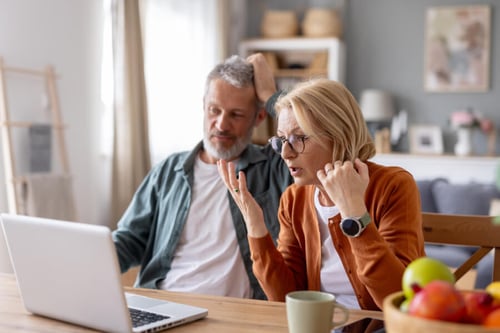 Deux seniors perplexes et inquiets consultent un site internet sur un ordinateur portable dans une cuisine moderne et lumineuse. L’homme, les mains sur la tête, semble stressé tandis que la femme, portant des lunettes, se tient la tempe en regardant l’écran avec une expression préoccupée. Cette scène illustre l’inquiétude liée à la découverte d’une arnaque en ligne, notamment l’arnaque aux loyers impayés ciblant les locataires. Les détails de leur environnement, comme les fruits sur la table et la décoration soignée, renforcent le réalisme de la situation et l’impact émotionnel de cette fraude financière sur des personnes vulnérables.