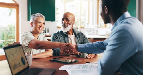 Scène chaleureuse montrant un agent immobilier accompagnant un couple de seniors dans le financement de leur projet immobilier. Autour d'une table en bois dans un cadre lumineux et accueillant, l'agent, un homme souriant en chemise bleue, serre la main du couple âgé pour sceller un accord. Le senior, barbu et vêtu d'une veste grise, et sa compagne aux cheveux gris clair, habillée d'un haut clair, affichent des expressions de satisfaction et de confiance. Sur la table, des documents financiers, un contrat immobilier et une tablette affichant des graphiques illustrent les différentes étapes de l'accompagnement personnalisé. Cette image reflète l'importance d'un soutien expert pour les seniors souhaitant concrétiser leur projet immobilier en toute sérénité.