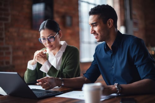 Deux professionnels discutent stratégiquement autour d'une table de travail dans un bureau moderne aux murs en briques apparentes. La femme, portant des lunettes et une veste verte sur une chemise blanche, consulte des documents tout en utilisant son ordinateur portable. L'homme, en chemise bleue, sourit en expliquant un point clé avec des gestes, illustrant un échange bienveillant et collaboratif. Cette scène représente un accompagnement personnalisé d’un vendeur à travers son cycle psychologique, mettant en avant l’écoute active, le mentorat et la formation continue pour optimiser ses performances commerciales et son bien-être professionnel. Idéal pour illustrer les méthodes d’accompagnement en vente et en management d’équipe.