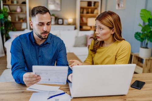 Deux personnes discutant de la renégociation d'un crédit immobilier autour d'une table en bois dans un salon chaleureux. Un homme en chemise bleue examine attentivement des documents papier, probablement un contrat de prêt immobilier, tandis qu'une femme aux cheveux mi-longs, vêtue d'un pull jaune, consulte des informations sur un ordinateur portable. Sur la table se trouvent également un stylo et un téléphone portable. L'arrière-plan montre une bibliothèque remplie de livres et des éléments de décoration, créant une ambiance propice à une discussion financière sérieuse et réfléchie. Cette scène illustre les étapes pratiques pour bien préparer la renégociation d'un prêt immobilier.