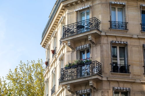 Découvrez un immeuble résidentiel typique en pierre avec balcons en fer forgé ornés de motifs élégants, situé en milieu urbain. Cet édifice haussmannien, aux fenêtres hautes et aux garde-corps travaillés, illustre parfaitement les logements concernés par les questions de congé pour reprise en droit immobilier. Les balcons sont agrémentés de jardinières fleuries, ajoutant une touche de verdure à l'architecture classique. Cette image évoque le cadre de vie souvent impacté par les procédures de congé pour reprise, notamment en cas de décès du bailleur, un sujet clé abordé dans notre article sur les droits des locataires et des propriétaires en matière de bail immobilier. Idéale pour accompagner des contenus sur la législation locative, les successions et les droits des héritiers dans le contexte d’un congé pour reprise.