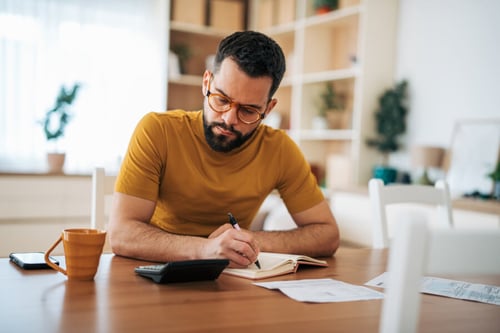 Homme concentré analysant des documents financiers et des taux de crédits immobiliers dans un bureau lumineux et moderne. Il prend des notes avec un stylo tout en consultant des papiers posés sur une table en bois, accompagné d'une calculatrice, d'un café et de relevés bancaires. Cette scène illustre l'étude des variations des taux de crédit immobilier influencées par le contexte économique international, idéal pour comprendre les impacts des marchés globaux sur les prêts immobiliers en France et en Europe.