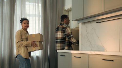 Deux personnes en train de déménager discutent dans une cuisine moderne après avoir déposé un préavis de départ. La femme, à gauche, tient un carton de déménagement marqué d’une étiquette tandis qu’elle semble poser une question ou échanger des informations. L’homme, à droite, place un autre carton sur un plan de travail de cuisine équipée de meubles clairs et d’un plan en marbre. Cette scène illustre une situation courante liée à la gestion d’un préavis de location et aux préparatifs de déménagement, notamment la possibilité de décaler la date de départ après le dépôt du préavis. Contexte idéal pour aborder les droits et démarches concernant la modification de la date de départ en fin de bail.