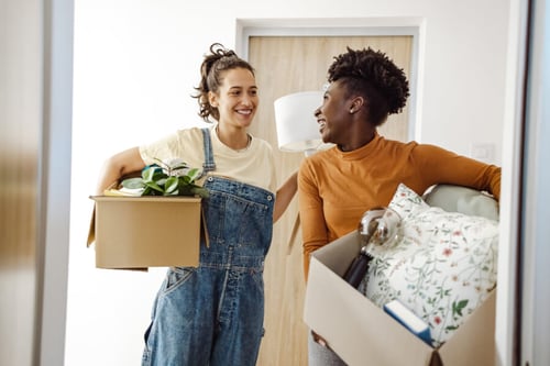 Deux personnes souriantes en train de préparer un déménagement facile et rapide, transportant des cartons dans un appartement lumineux. La femme à gauche porte une salopette en jean et un t-shirt blanc, tenant un carton rempli de fournitures et d’une plante verte. L’homme à droite, vêtu d’un t-shirt orange, porte un carton contenant des coussins décoratifs et des objets ménagers. Ils semblent organiser efficacement leurs affaires pour optimiser le temps et l’espace, illustrant des astuces pratiques pour un déménagement sans stress. Ambiance positive et collaborative dans un cadre intérieur moderne. Idéal pour des conseils de déménagement organisés et sereins.