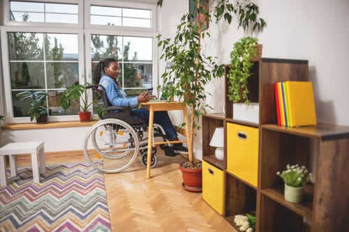 Femme en fauteuil roulant travaillant sur un ordinateur portable dans un salon lumineux et accessible. Le logement adapté comprend des fenêtres larges laissant entrer la lumière naturelle, des étagères à hauteur accessible, et des plantes décoratives disposées sur des meubles ergonomiques. Le sol plat et sans obstacles ainsi que le large espace de circulation illustrent les aménagements nécessaires pour les locataires handicapés. Cette image met en avant l'importance de l'accessibilité et des droits des locataires en situation de handicap dans un logement locatif, conformément aux normes d'aménagement et de non-discrimination en vigueur. Idéale pour illustrer un article sur les droits des locataires handicapés et les adaptations nécessaires du logement.