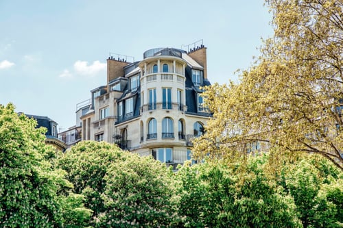 Découvrez un exemple typique de l'immobilier résidentiel en France illustrant la hausse des loyers analysée par l'Observatoire 2026. Cette photographie montre un immeuble haussmannien élégant à plusieurs étages, situé dans un quartier urbain probablement parisien. Les façades en pierre de taille, les balcons en fer forgé et les fenêtres hautes témoignent du charme architectural recherché, souvent associé à des loyers élevés. En premier plan, une végétation dense avec des arbres en fleurs souligne l’attrait des espaces verts dans les zones résidentielles prisées. Ce type de bien, emblématique des grandes villes françaises, est directement concerné par l’évolution des prix du marché locatif, un sujet central abordé dans notre analyse sur la hausse des loyers en France selon les dernières données de l'Observatoire 2026.