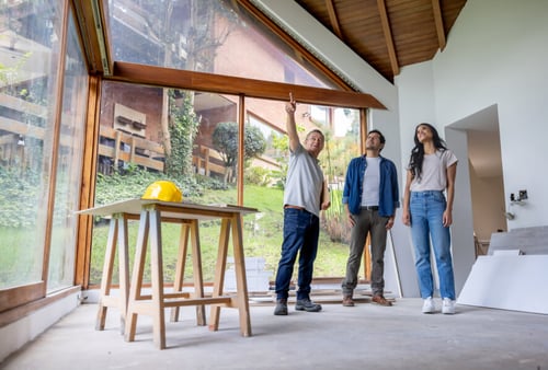 Trois personnes discutent d’un projet de construction ou de rénovation dans une pièce lumineuse avec de grandes baies vitrées offrant une vue sur un jardin extérieur. Un homme en t-shirt gris pointe vers le plafond, probablement pour évaluer les travaux à réaliser, tandis qu’un autre homme en veste bleue et une femme en t-shirt blanc et jeans l’écoutent attentivement. Au centre de la pièce se trouve un petit chevalet de travail en bois avec un casque de chantier jaune posé dessus, suggérant une préparation pour des travaux de construction ou d’aménagement. Cette scène illustre une étape clé dans l’évaluation d’un investissement locatif, notamment l’analyse des coûts et des aménagements nécessaires pour optimiser la rentabilité d’un bien immobilier neuf ou rénové. Idéale pour aborder les avantages de faire construire dans le cadre d’un projet locatif, cette image met en avant la planification et la collaboration entre investisseurs et professionnels du bâtiment.