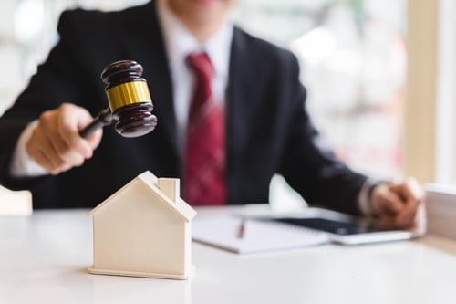 Juge en robe noire avec cravate rouge tenant un marteau de justice posé sur une maquette de maison blanche symbolisant une vente immobilière aux enchères judiciaires. Contexte de saisie immobilière et d'achat de bien saisi par décision de justice, illustrant les opportunités et procédures liées à l'acquisition d'un logement saisi à prix potentiellement avantageux. Idéal pour un article sur les avantages et précautions à prendre lors de l'achat d'un bien immobilier saisi.