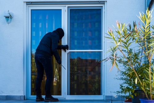 Homme en tenue sombre tentant de s'introduire par une fenêtre entrouverte d'une maison en plein jour. La scène illustre un cambriolage par effraction via une fenêtre mal fermée, un risque accru pour les propriétaires en matière de couverture d'assurance habitation. La maison, peinte en blanc avec des cadres de fenêtres bleus, montre l'importance de sécuriser les accès pour éviter les refus d'indemnisation par les assureurs en cas de négligence comme des fenêtres laissées ouvertes. Ce visuel met en lumière les conséquences potentielles d'une intrusion sur les contrats d'assurance et les bonnes pratiques pour protéger son domicile.