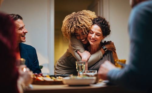 Groupe d’amis ou famille réunis autour d’un repas convivial dans un restaurant, illustrant l’ambiance chaleureuse d’un hébergement à titre gratuit entre proches. Deux femmes s’étreignent joyeusement tandis qu’un homme sourit en arrière-plan, symbolisant les relations de confiance et les règles à respecter pour accueillir un proche en location sans loyer. Cette scène évoque les aspects légaux, fiscaux et pratiques abordés dans l’article sur l’hébergement gratuit en logement loué, comme la déclaration aux impôts, l’accord du propriétaire et la durée du séjour. Idéale pour accompagner un contenu sur les obligations et conseils pour héberger un ami ou un membre de la famille en toute sérénité.