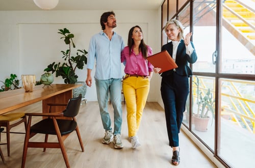 Trois personnes discutent d’un projet immobilier dans un appartement lumineux et moderne. À gauche, un homme barbu en chemise bleue et jean observe les lieux avec intérêt. Au centre, une femme en haut rose et pantalon jaune écoute attentivement, tandis qu’à droite, une agente immobilière souriante, vêtue d’un tailleur bleu, présente les détails d’un investissement en tenant un dossier rouge. L’espace est décoré avec des plantes vertes et un mobilier design, offrant une ambiance chaleureuse et professionnelle. Cette scène illustre les options d’investissement immobilier, qu’il s’agisse d’acheter un bien en direct ou de passer par une SCPI pour diversifier son portefeuille. Idéal pour comprendre les avantages et inconvénients de chaque solution.
