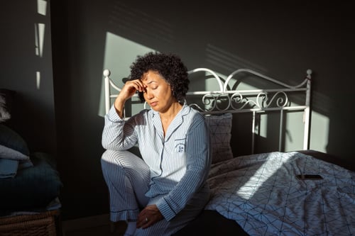 Femme stressée en pyjama rayé assise sur le bord d’un lit dans une chambre mal insonorisée, se tenant la tête dans les mains. La lumière naturelle filtre à travers une fenêtre partiellement ouverte, révélant un environnement intérieur calme mais suggérant des nuisances sonores extérieures. Le désordre des draps et l’expression de fatigue illustrent l’impact d’un logement bruyant sur la qualité de vie et le bien-être. Cette image évoque les problèmes courants de location liés à une mauvaise isolation phonique et renvoie aux solutions juridiques et pratiques pour les locataires confrontés à ce type de désagrément, comme détaillé dans l’article Quels recours si vous louez un logement mal insonorisé.