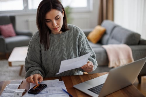 Femme analysant des documents liés à un loyer impayé avec un ordinateur portable et une calculatrice sur une table de salon. Illustration des nouvelles règles pour les locataires concernant les loyers impayés à partir de janvier 2027. La personne semble étudier des relevés bancaires et des contrats de location tout en vérifiant des calculs financiers, reflétant les démarches administratives et juridiques en cas de difficultés de paiement du loyer. Contexte de gestion locative et de protection des droits des locataires.