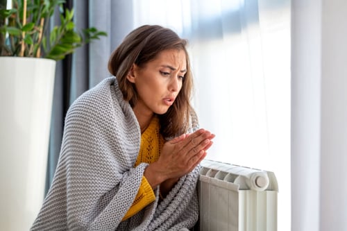 Femme enveloppée dans une couverture se tenant la poitrine près d’un radiateur froid lors d’une panne de chauffage en intérieur