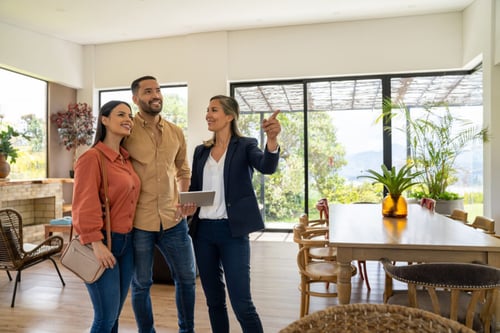 Couple visitant une maison contemporaine avec une agente immobilière pour illustrer l'impact de la proposition de loi Létard sur les agences immobilières. La scène montre une discussion professionnelle dans un cadre lumineux et moderne, mettant en avant les échanges entre clients et agent. Le salon spacieux avec grandes baies vitrées, mobilier design et décoration soignée reflète un bien immobilier haut de gamme. Cette image s’intègre dans un article expliquant les nouvelles obligations et changements pour les agences immobilières suite à la proposition de loi Létard, notamment en matière de transparence, de formation des agents et de protection des consommateurs. Idéale pour évoquer les bonnes pratiques et l’accompagnement personnalisé dans le secteur immobilier.