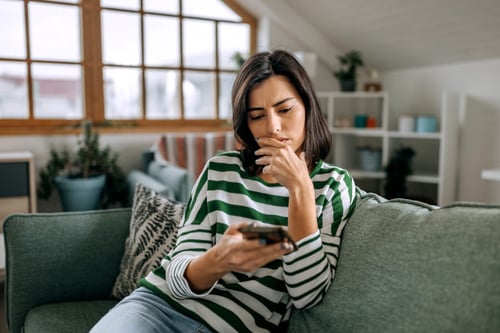 Femme inquiète assise sur un canapé vert dans un salon lumineux avec grandes fenêtres, consultant son téléphone portable avec un air préoccupé. Elle semble chercher des solutions en ligne concernant un propriétaire non réactif ou absent. Contexte illustrant les démarches à suivre lorsque un propriétaire ne répond plus aux locataires, incluant recherche d’informations juridiques ou conseils pratiques sur internet. Intérieur moderne et chaleureux avec étagères et plantes en arrière-plan. Idéal pour un article sur les recours en cas de silence du propriétaire.
