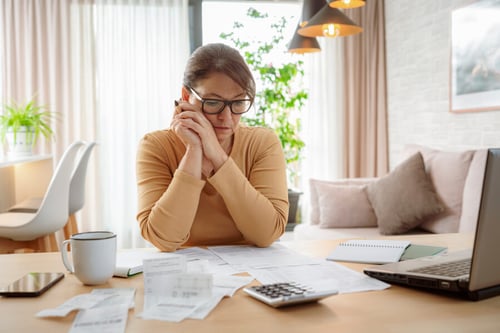 Femme stressée en train de gérer une dette locative et des problèmes de surendettement à domicile. Elle consulte des documents financiers, factures et relevés bancaires étalés sur une table en bois avec un ordinateur portable, une calculatrice et une tasse de café. La scène se déroule dans un salon lumineux et cosy, illustrant les difficultés financières et l'impact du surendettement sur les propriétaires. Cette image reflète les défis liés à la gestion des dettes locatives et les conséquences sur la situation personnelle et financière des propriétaires bailleurs. Idéale pour accompagner un article sur les solutions et recours en cas de surendettement immobilier.