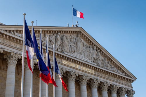 Bâtiment de l'Assemblée Nationale française avec drapeaux flottants sous un ciel bleu clair. Cette image illustre le siège du pouvoir législatif en France où se discute actuellement une mission gouvernementale sur les impayés de loyers pour les propriétaires. La façade néoclassique ornée de colonnes et de sculptures représente l'institution chargée d'examiner les nouvelles mesures et réglementations visant à soutenir les propriétaires face aux difficultés de recouvrement des loyers impayés. Contexte politique et législatif lié aux réformes du logement en France. Idéal pour un article sur les droits des propriétaires et les solutions aux impayés de loyer.