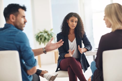 Une femme professionnelle d’origine africaine en réunion tendue avec deux collègues dans un bureau lumineux, exprimant son mécontentement face à une situation de discrimination. Elle est assise sur une chaise blanche, vêtue d’un blazer bleu marine, d’un t-shirt gris et d’un pantalon bordeaux, gesticulant pour appuyer ses propos. À gauche, un homme en chemise bleue semble réagir avec désapprobation ou surprise, tandis qu’à droite, une femme blonde en pull noir observe la scène avec attention. Cette image illustre un échange lié à des pratiques discriminatoires, notamment dans le cadre d’une recherche de logement et des droits des locataires face à un bailleur raciste. Contexte adapté pour un article sur les recours contre la discrimination raciale dans l’immobilier.