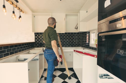 Homme en train de rénover une cuisine à moindre coût en posant un niveau à bulle sur un meuble pour vérifier l'alignement lors de travaux de bricolage. La cuisine moderne présente des carreaux de faïence noirs et blancs, un plan de travail partiellement installé, et des outils de mesure et de fixation posés à proximité. Ambiance lumineuse grâce à un éclairage suspendu design et des murs clairs. Idéal pour des conseils pratiques sur la rénovation de cuisine sans gros budget ni intervention du bailleur, en utilisant des techniques simples et des outils accessibles. Astuces pour optimiser l’espace et améliorer l’esthétique sans engager de dépenses importantes.
