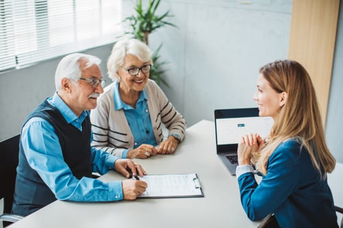 Couple de seniors souriants signant un contrat de tontine avec une conseillère financière dans un bureau lumineux. La scène illustre une discussion autour des avantages de la tontine pour les propriétaires souhaitant sécuriser la transmission de leur bien immobilier. La conseillère, équipée d’un ordinateur portable, explique les détails du document tout en offrant un accompagnement personnalisé. Idéal pour comprendre les mécanismes juridiques et fiscaux de la tontine en France et optimiser la gestion de son patrimoine immobilier. Image représentative des solutions de prévoyance pour les seniors et les couples propriétaires.