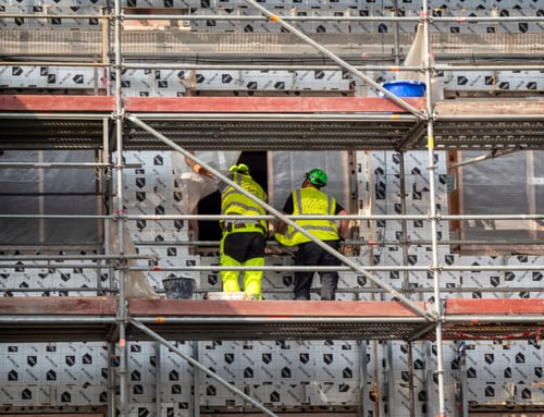 Trois ouvriers en tenue de sécurité jaune et casques verts travaillent sur un échafaudage installé devant la façade d’un immeuble en rénovation. Les travaux semblent concerner l’isolation extérieure, avec des panneaux isolants déjà posés partiellement et des structures métalliques de soutien visibles. La scène illustre des travaux de rénovation énergétique ou de ravalement de façade, pouvant entraîner des nuisances pour les locataires et justifier une demande de réduction de loyer en raison des désagréments subis. Ce type de chantier est souvent évoqué dans les discussions sur les droits des locataires face aux perturbations liées aux améliorations du bâtiment.