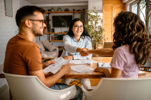 Trois personnes souriantes en pleine signature d'un mandat de représentation immobilier autour d'une table en bois dans un salon lumineux : une agente immobilière en chemise claire et lunettes, serrant la main d'une cliente aux cheveux longs et bouclés, tandis qu'un homme en pull orange consulte des documents posés devant eux, illustrant ainsi la confiance et le professionnalisme dans les transactions immobilières sécurisées.