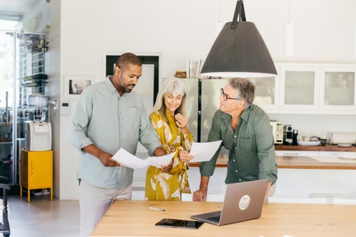 Trois personnes discutant autour d’une table dans une cuisine moderne et lumineuse pour analyser des documents liés à la gestion locative. Une femme aux cheveux gris vêtue d’une robe jaune, un homme en chemise vert clair et un autre homme en chemise verte avec des lunettes examinent attentivement des feuilles imprimées, tandis qu’un ordinateur portable ouvert affiche probablement des informations complémentaires. Scène idéale illustrant une réunion collaborative pour la préparation ou le suivi d’un bail mobilité, dans un cadre domestique propice aux échanges professionnels.