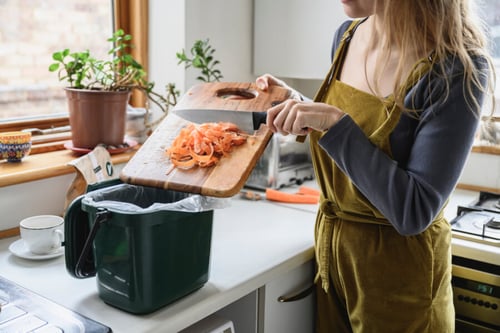Une personne en tablier vert préparant des épluchures de carottes dans une cuisine bien éclairée et les déposant dans un seau de compost vert équipé d’un couvercle et d’un sac compostable, illustration concrète des gestes écologiques de compostage en intérieur adaptés aux logements en location.
