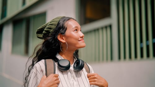 Jeune femme souriante et détendue en extérieur devant un bâtiment résidentiel avec des fenêtres vert pâle, portant des écouteurs autour du cou, un beanie vert et une veste striée, tenant les bretelles de son sac à dos, symbolisant la recherche de logement sereinement pendant une période d’essai professionnelle dans une nouvelle ville.