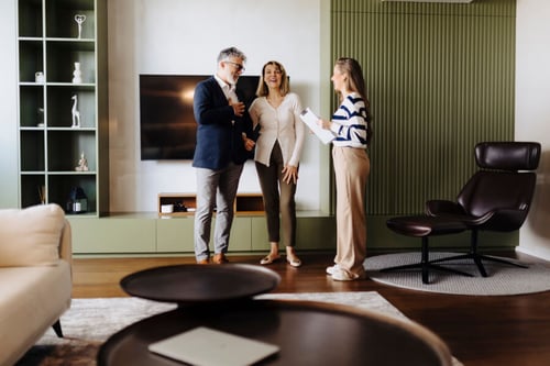 Une gestionnaire locative souriante présentant un document à un couple de propriétaires expatriés dans un salon moderne et lumineux, équipé d’un canapé beige, d’une table basse en bois, d’un fauteuil design et d’une télévision murale, illustrant un accompagnement personnalisé pour la gestion de leur bien immobilier pendant leur expatriation.