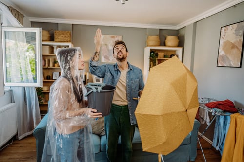 Un locataire et une locataire en situation de stress dans un salon après un dégât des eaux : la femme, vêtue d'une housse de protection transparente, tient un seau pour récupérer l'eau, tandis que l'homme, en chemise denim et pantalon vert, lève la main en signe d'impuissance ou d'incompréhension devant une flaque d'eau, avec un parapluie ouvert à côté de lui pour tenter de limiter les dégâts. L'arrière-plan montre un intérieur moderne avec des meubles et des accessoires de décoration, illustrant les conséquences immédiates d'une fuite ou d'une infiltration d'eau dans un logement.