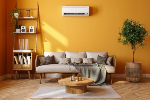 Découvrez un intérieur meublé moderne et chaleureux idéal pour comprendre l’encadrement des loyers des locations meublées. Ce salon lumineux aux murs orange clair est équipé d’un canapé confortable en tissu beige avec plusieurs coussins assortis, d’une couverture grise et d’une table basse en bois circulaire. À gauche, une étagère en bois élégamment organisée avec des plantes vertes, des livres, des enceintes et des éléments décoratifs. À droite, un grand pot en osier abrite une plante verte, ajoutant une touche naturelle à l’espace. Au-dessus du canapé, un climatiseur mural discret assure le confort thermique. Ce cadre illustre parfaitement les critères de décoration et d’équipement nécessaires pour une location meublée conforme aux réglementations sur l’encadrement des loyers. Parfait pour un logement étudiant ou une résidence temporaire, cet aménagement répond aux exigences légales tout en offrant un environnement accueillant.