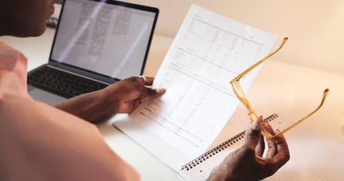 Une personne analysant attentivement des documents administratifs relatifs à des procédures financières ou juridiques, avec un ordinateur portable ouvert affichant du texte, tout en tenant une paire de lunettes ; illustration concrète pour un article sur les nouveautés concernant la saisie sur salaire pour loyers impayés, mettant en avant la vérification et l'étude de détails réglementaires.