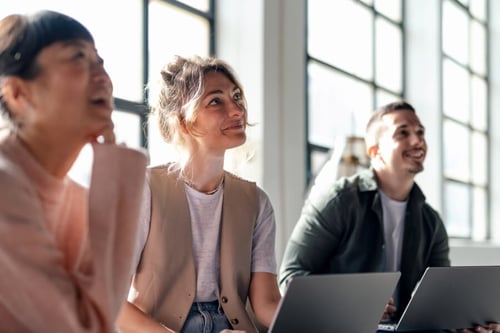 Trois professionnels de l'immobilier participent activement à une formation en équipe dans un espace lumineux et moderne. La session semble se dérouler dans un bureau avec de grandes fenêtres laissant entrer la lumière naturelle. Une femme au centre, souriante et attentive, porte une veste beige sur un t-shirt gris et discute avec ses collègues. À gauche, un homme en arrière-plan écoute avec intérêt, tandis qu’à droite, un autre collaborateur, également souriant, travaille sur son ordinateur portable. Cette image illustre parfaitement l’engagement et la dynamique d’une formation des collaborateurs immobiliers prévue pour juillet 2026, mettant en avant l’importance de la collaboration et de l’apprentissage continu dans le secteur immobilier.