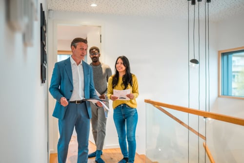 Un agent immobilier en costume bleu marine discutant avec deux jeunes investisseurs dans un intérieur moderne et lumineux, probablement un bien en visite pour un futur investissement locatif. L’homme, à gauche, tient des documents tout en expliquant des détails, tandis que les deux investisseurs, une femme en pull jaune et jeans bleus ainsi qu’un homme en pull gris, écoutent attentivement avec des documents à la main, illustrant ainsi les nouveaux profils d’acheteurs intéressés par l’investissement locatif. La scène se déroule près d’un escalier avec une rampe en verre, dans un espace épuré et contemporain.