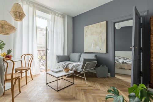 Description SEO optimisée pour texte alternatif : intérieur moderne d'un studio en location meublée éligible à la Cotisation Foncière des Entreprises (CFE) avec salon ouvert sur balcon. Pièce lumineuse avec sol en parquet clair en motif chevron, murs gris anthracite et grande baie vitrée donnant sur un petit balcon. Mobilier fonctionnel incluant un canapé-lit gris avec coussins, une table basse blanche à structure métallique, et deux chaises en bois style bistrot autour d'une table ronde. Éclairage design avec suspension en osier naturel et lampe murale. Espace optimisé avec porte ouverte sur une chambre équipée d'un lit et rangements. Décoration épurée avec une grande toile abstraite au mur et plantes vertes pour une ambiance chaleureuse. Idéal pour illustrer les spécificités des logements meublés soumis à la CFE.