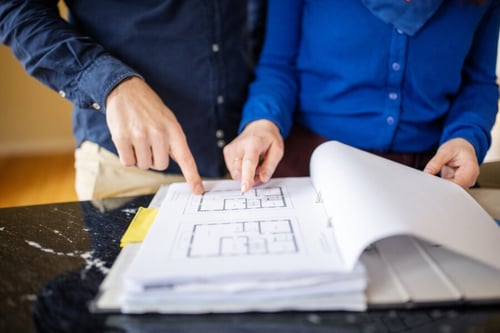 Deux personnes étudiant attentivement un plan d'appartement avec un stylo jaune pour marquer des annotations. Les individus, vêtus de chemises bleues, semblent discuter des avantages et inconvénients des différentes configurations de logement, notamment entre un rez-de-chaussée et un étage. Cette scène illustre la phase de recherche et de comparaison essentielle lors de la location d'un appartement, mettant en avant l'importance de bien analyser les plans pour faire un choix éclairé. Idéal pour accompagner un article sur les critères de sélection d'un logement en fonction de son niveau.