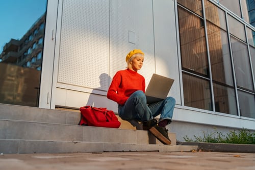 Étudiante assise sur les marches d’un bâtiment moderne en train de consulter son ordinateur portable, probablement à la recherche d’un logement étudiant. Elle porte un pull rouge chaud, un jean bleu et un béret jaune, avec un sac rouge posé à côté d’elle. Scène en extérieur avec un éclairage naturel de fin de journée, illustrant une ambiance de préparation et d’organisation pour les démarches liées à la vie étudiante en ville.