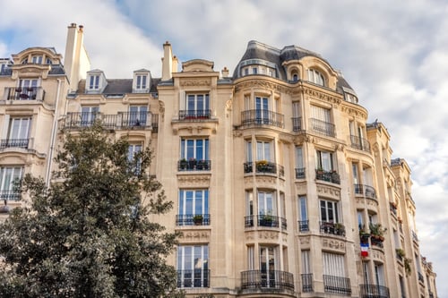 Immeuble parisien typique avec architecture haussmannienne de plusieurs étages, présentant des balcons en fer forgé, des fenêtres à petits carreaux et des façades ornées de moulures, sous un ciel partiellement nuageux, illustrant le type de bâtiment concerné par les réglementations immobilières en vigueur, dont la loi ALUR 2025 imposant une formation obligatoire pour les professionnels du secteur.