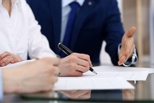 Deux personnes en train de signer un contrat immobilier ou professionnel avec un agent en costume : une personne remplit et paraphe le document avec un stylo tandis qu'un professionnel en costume bleu, probablement un agent mandaté, tend la main pour finaliser l'accord, illustrant ainsi les interactions possibles dans le cadre d'un mandat d’entremise ou de représentation.