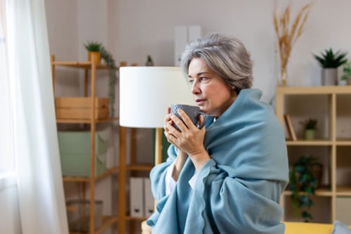 Femme âgée enveloppée dans une couverture bleue pour se protéger du froid dans un logement mal isolé. Elle tient une tasse chaude entre ses mains pour se réchauffer, illustrant les difficultés rencontrées lors d’une vague de froid dans une maison peu chauffée. À l’arrière-plan, un intérieur cosy avec des étagères en bois, des plantes vertes et une lumière douce, mettant en évidence le contraste entre le confort apparent et la sensation de froid. Image adaptée pour un article sur les solutions pour améliorer le quotidien en période de grand froid dans un habitat mal isolé.