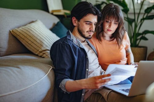 Jeune couple assis sur un canapé en train de consulter attentivement des documents papier et un ordinateur portable, probablement liés à leur situation locative, avec une expression concentrée et sérieuse, dans un intérieur cosy avec des coussins décoratifs et des plantes vertes en arrière-plan, illustrant une recherche d'informations sur les droits des locataires lorsque le propriétaire vend le logement en cours de bail.