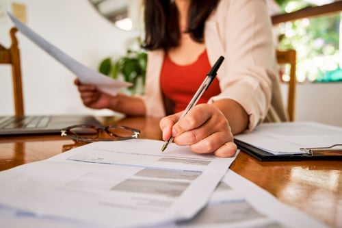 Femme en train de remplir un document administratif lié à la location d'un logement, assise à une table avec un stylo à la main, entourée de formulaires et d'un clipboard, illustrant les démarches pour déterminer le montant de la caution locative. Un ordinateur portable et des lunettes sont également visibles, soulignant l'importance de la préparation et de la vérification des informations financières.
