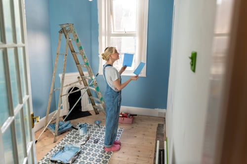 Jeune femme en tenue de bricolage comparant des échantillons de couleur bleue contre un mur fraîchement peint dans une pièce en rénovation, debout sur un sol en bois partiellement protégé par une bâche et des feuilles de protection, avec une échelle et des outils de peinture à proximité, illustrant les travaux de peinture intérieure souvent nécessaires lors d'un état des lieux.